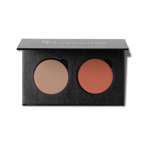 An open black makeup palette with two round pressed powder pans, one light beige and one warm terracotta orange, on a white background. The brand name 'B:Undeniable' is printed on the lid.