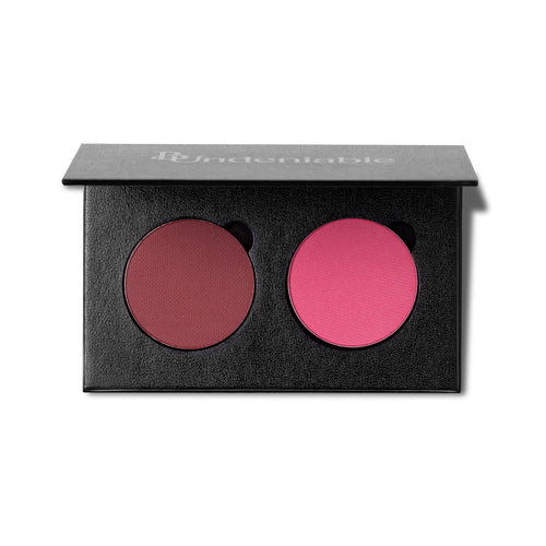 An open black makeup palette with two round pressed powder pans, one muted berry red and one bright pink, on a white background. The brand name 'B:Undeniable' is printed in white on the lid.