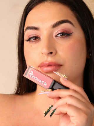 Woman holding a pink lip gloss with 'bundeniable' branding.